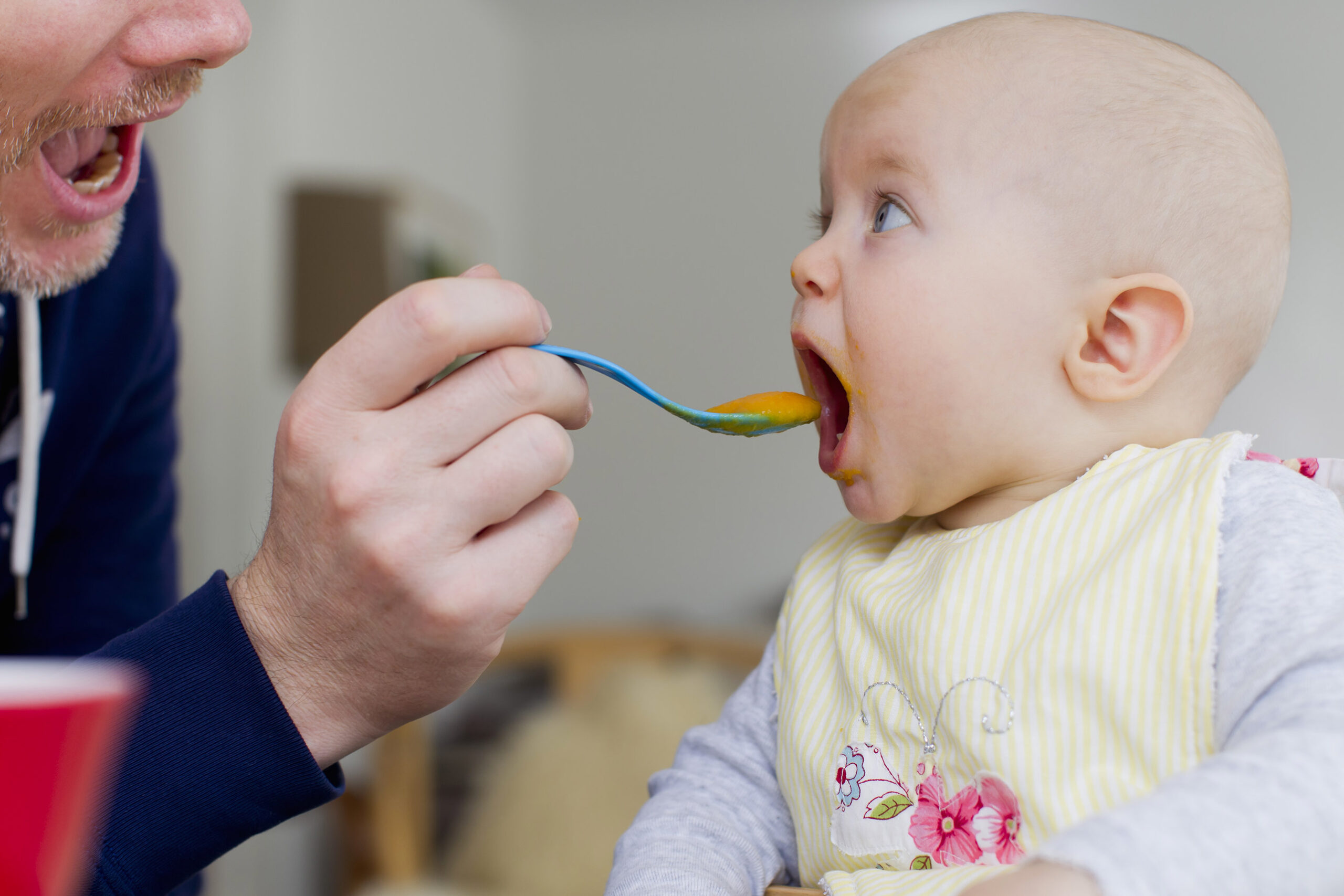 a close up photo of a father feeding his baby with a spoon. The baby has their mouth wide open ready to eat the food on the spoon.